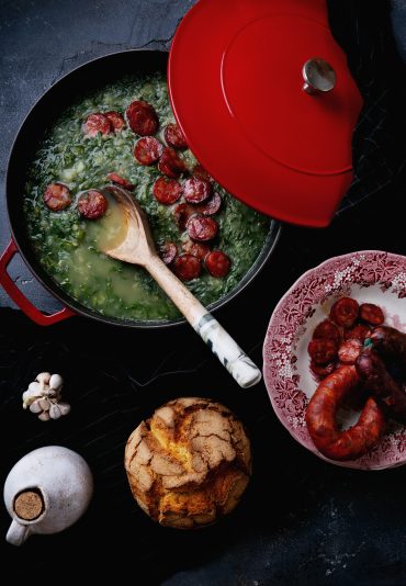 Caldo Verde. Traditional Portuguese Soup Caldo Verde Soup with greens and chopped chorizo on the top in the cooking pan with wooden spoon. Traditional round bread and chorizo on the plate. Top View