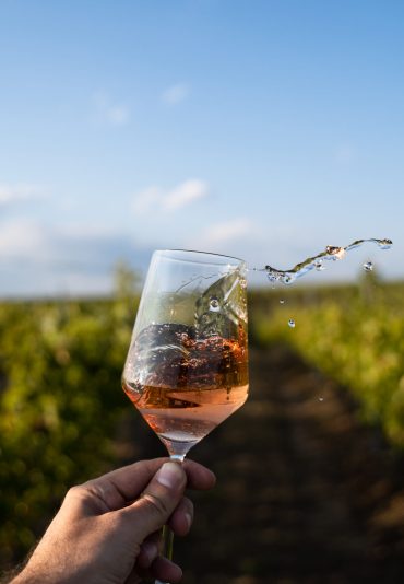 perspective point of view closeup of Caucasian male hand agitating and spilling with a splash glass of rose wine in the morning light with green vineyard and blue sky in the background