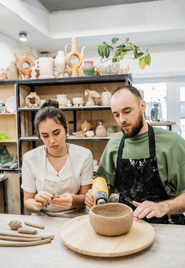 Craftsman drying earthenware with heat gun while girlfriend shaping clay in ceramic workshop