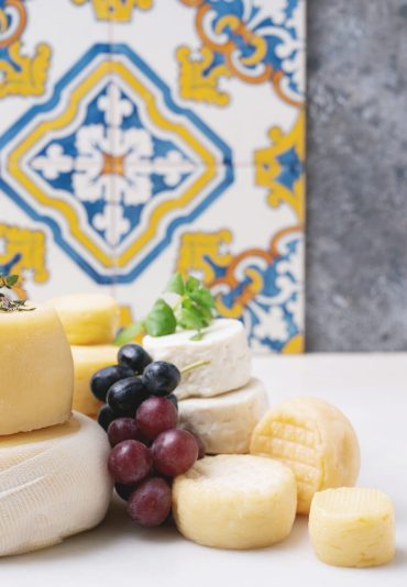 Traditional Portuguese semi-soft cheeses from evora alentejo and azores regions on the tray, served with fresh grapes, honey and herbs. Side view. Selective focus. Azulejo pattern.